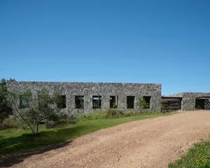 A modern building constructed of stone with a dirt driveway and greenery.