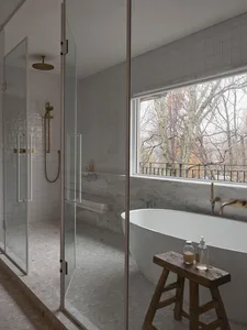 Modern minimalist bathroom featuring a white marble tub, glass shower enclosure, and a window overlooking trees.