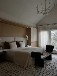 A modern bedroom with a bed dressed in textured, neutral-colored linens and a minimalist black bench against a textured rug. 