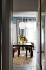 A modern dining room with a wooden table and leather chairs, featuring a white pendant light and sheer curtains.