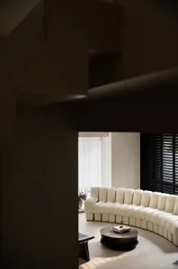 A modern living room featuring a circular sofa with plush cushions, a round coffee table, and black window shutters. The spac