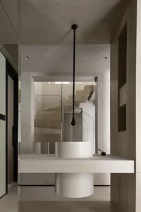 A modern minimalist bathroom with a white floating sink and a glass-enclosed staircase in the background.