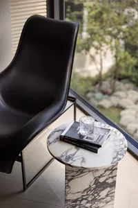 A minimalist modern penthouse with a sleek black chair, marble side table, and a glass of water, featuring a view of greenery