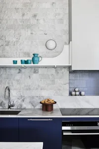 A modern kitchen with marble countertops and a blue cabinet beneath, featuring a marble backsplash, blue cups on a shelf, and