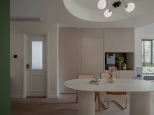 A modern dining room featuring a white marble table with wooden legs and chairs, next to a kitchen area with white cabinets a