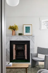 A modern living room with marble fireplace, grey armchairs, and green vase on mantel.