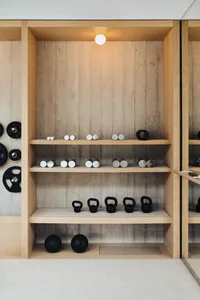 A minimalist fitness room with concrete walls and wooden shelves, featuring dumbbells, kettlebells, and medicine balls.
