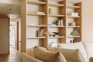 A minimalist living room features a wooden shelf with books, a white floor lamp, and a beige sofa.