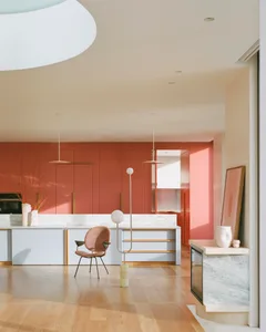A modern kitchen with white marble countertops and cabinets, featuring a dining table, pendant lights, and a pink chair.