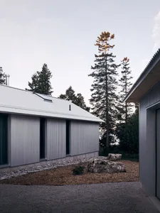 A minimalist building with a metal exterior features a sloped roof and large windows, surrounded by a gravel walkway and a ro