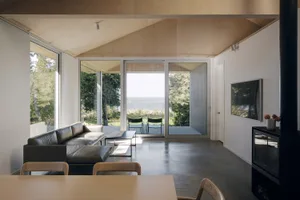A minimalist living room with concrete flooring, a large leather sofa, and a dining area with chairs and a table.
