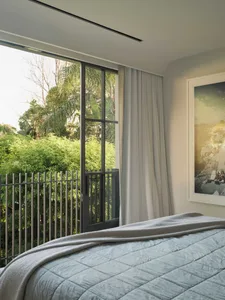 A bedroom with a balcony overlooking lush green plants, featuring a bed with grey bedding, and light grey curtains.