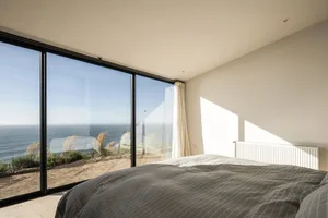 A modern bedroom with a large window overlooking the sea, featuring a bed with striped bedding and a white radiator.
