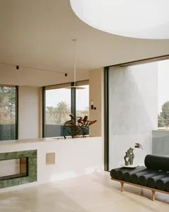 A modern minimalist living room with large windows, a green marble fireplace, and a black bench with cylindrical cushions.