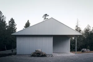A minimalist building with a metal cladding exterior, featuring an open garage door and surrounded by trees and rocks.