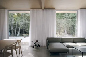 A modern, minimalist living room with large windows, white curtains, and a comfortable sofa in front of a concrete floor.