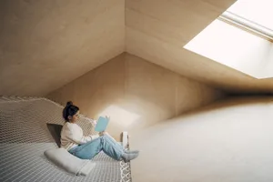 A woman sits on a hammock in an attic space with a skylight window.