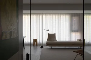 A minimalist residential lounge with a beige chaise lounge, a floor lamp, and sheer curtains allowing light to filter in.