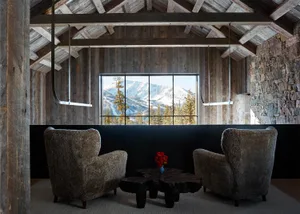 A modern living room with a wooden interior, featuring a coffee table, armchairs, and a large window with a mountain view.