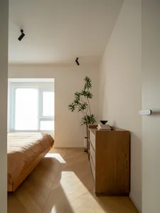 A minimalist bedroom featuring a bed with wood frame and beige bedding, surrounded by concrete walls and a wooden dresser.
