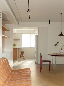 A minimalist white space featuring a wooden bench, a red chair, a round wooden table, and a wooden counter with a stool and a