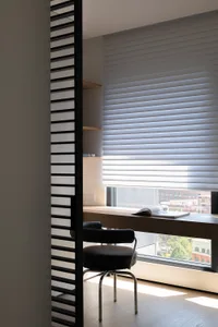 A minimalist home office with a wooden desk, a black chair, and a window with blinds.