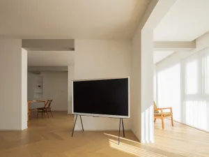 A minimalist living room with a large black screen, wooden flooring, and a window with white blinds.