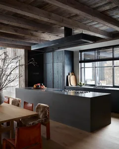 A modern minimalist kitchen featuring black cabinets, wood beams, and a dining table with a fur throw.