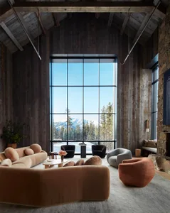 Modern living room with large windows overlooking snowy mountains, featuring a comfortable sofa, armchairs, and a stone firep