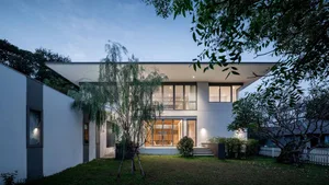 A modern residential building with a white exterior and large windows at dusk, surrounded by greenery and trees.