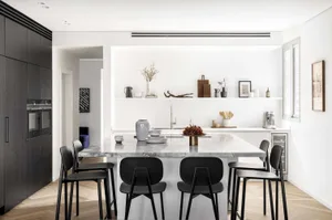 A modern minimalist kitchen with a marble island and black stools. The kitchen is well-equipped with stainless steel applianc