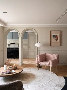 A minimalist living room featuring a pink upholstered chair, a round wooden coffee table, and a circular rug on a wooden floo