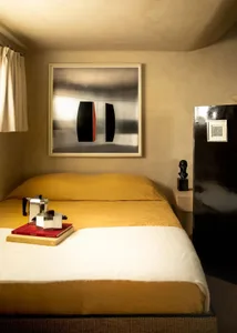 A modern bedroom with a yellow linen bed featuring a painting, a sculpture, and a coffee maker.