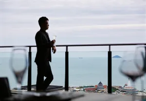Man standing on a balcony overlooking the ocean with a glass of wine.