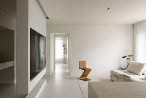 Modern minimalist living room featuring a white sofa, a sleek wooden chair, and a flat-screen TV mounted on a concrete wall.