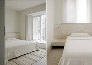 A minimalist bedroom with a stone headboard, white bedding, and a window with blinds.