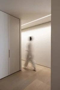 A blurred image of a person walking through a minimalist residential hallway with wooden flooring and white walls.