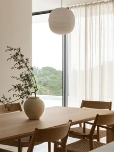 A modern minimalist dining room with a wooden table, chairs, and a vase with plant branches on it, near a large window with w