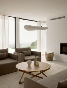 A modern minimalist living room featuring beige upholstered sofas, a round wooden coffee table, and a hanging lamp with a whi