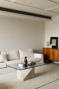 A modern living room with minimalist furniture and neutral-toned decor, featuring a glass-top coffee table, a wooden sideboar