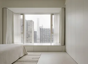 A minimalist bedroom with a bed, a rug, and a large window overlooking a cityscape.
