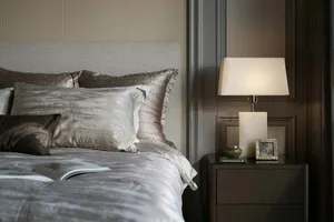 A neatly made bed adorned with luxury bedding, set against a muted backdrop, illuminated by a warm-toned lamp on a wooden nig