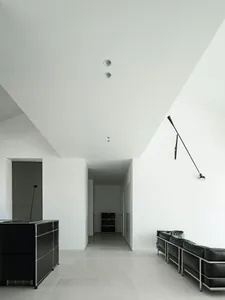 A minimalist interior space featuring a concrete floor with a long, sleek, black floor lamp and a black sofa with metal legs.