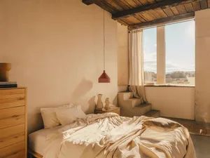 Minimalist bedroom with light-colored bedding and natural fiber textiles, featuring a red pendant light and a large window wi