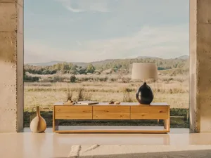 Minimalist interior design featuring wooden furniture, a lamp, and a vase in a spacious, open-concept room with a view of hil