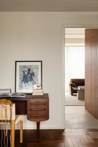 A minimalist workspace featuring a wooden desk and chair, with a framed artwork and books on the desk.