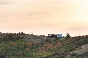 A modern house with steel and glass walls set in a hilly landscape during sunset.