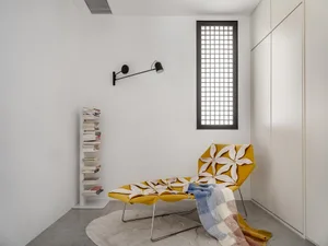 A minimalist bedroom featuring a yellow chaise lounge with flower upholstery, a blue and white striped blanket, and a bookshe