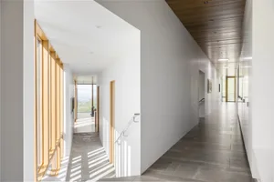 A minimalist hallway with white walls, wooden ceiling, concrete flooring, and large windows providing natural light.