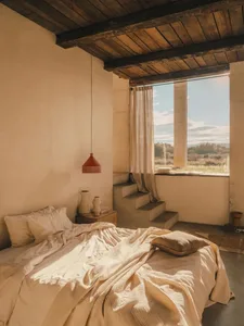 A minimalist bedroom featuring a bed with concrete mattress and beige bedding under a hanging red pendant light near a window